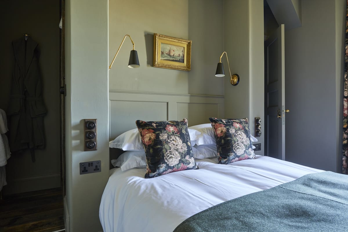 A neatly made bed with floral pillows, a framed picture on the wall, and a coat rack with a robe hanging.