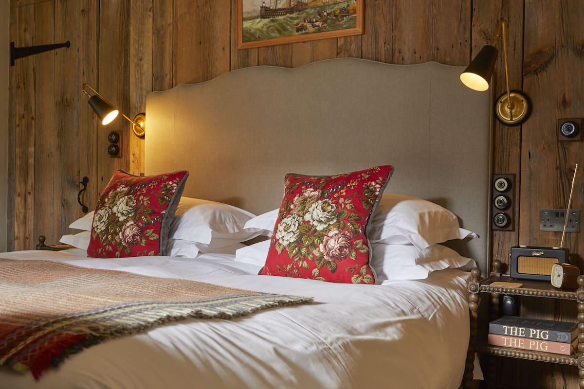 A neatly made bed with white linens and red floral pillows, a wooden headboard, and a nightstand with a radio and books.
