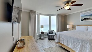 A hotel room with a bed, a desk, a chair, a ceiling fan, and a view of the sea. - Emerald Beach #830 | Stunning View, Complimentary Seasonal Beach Service (Panama City Beach)