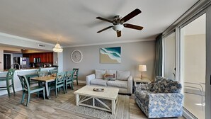 A spacious living area with a dining table, chairs, a sofa, a patterned armchair, a ceiling fan, a kitchen in the background, and a large window providing natural light. - Emerald Beach #830 | Stunning View, Complimentary Seasonal Beach Service (Panama City Beach)