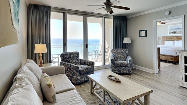 A hotel room with a sofa, two patterned armchairs, a coffee table, a ceiling fan, and a view of the ocean through large windows. - Emerald Beach #830 | Stunning View, Complimentary Seasonal Beach Service (Panama City Beach)