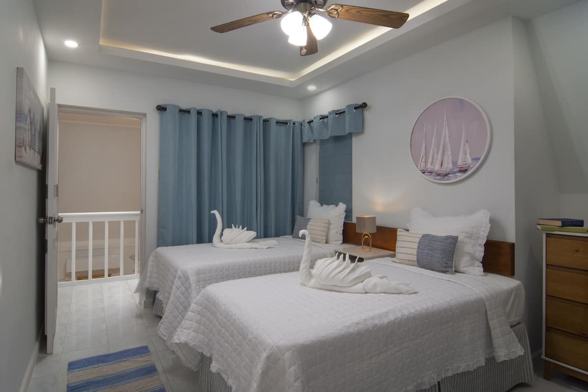 A hotel room with two beds, a ceiling fan, a painting of sailboats, and a view of the sea through the window.
