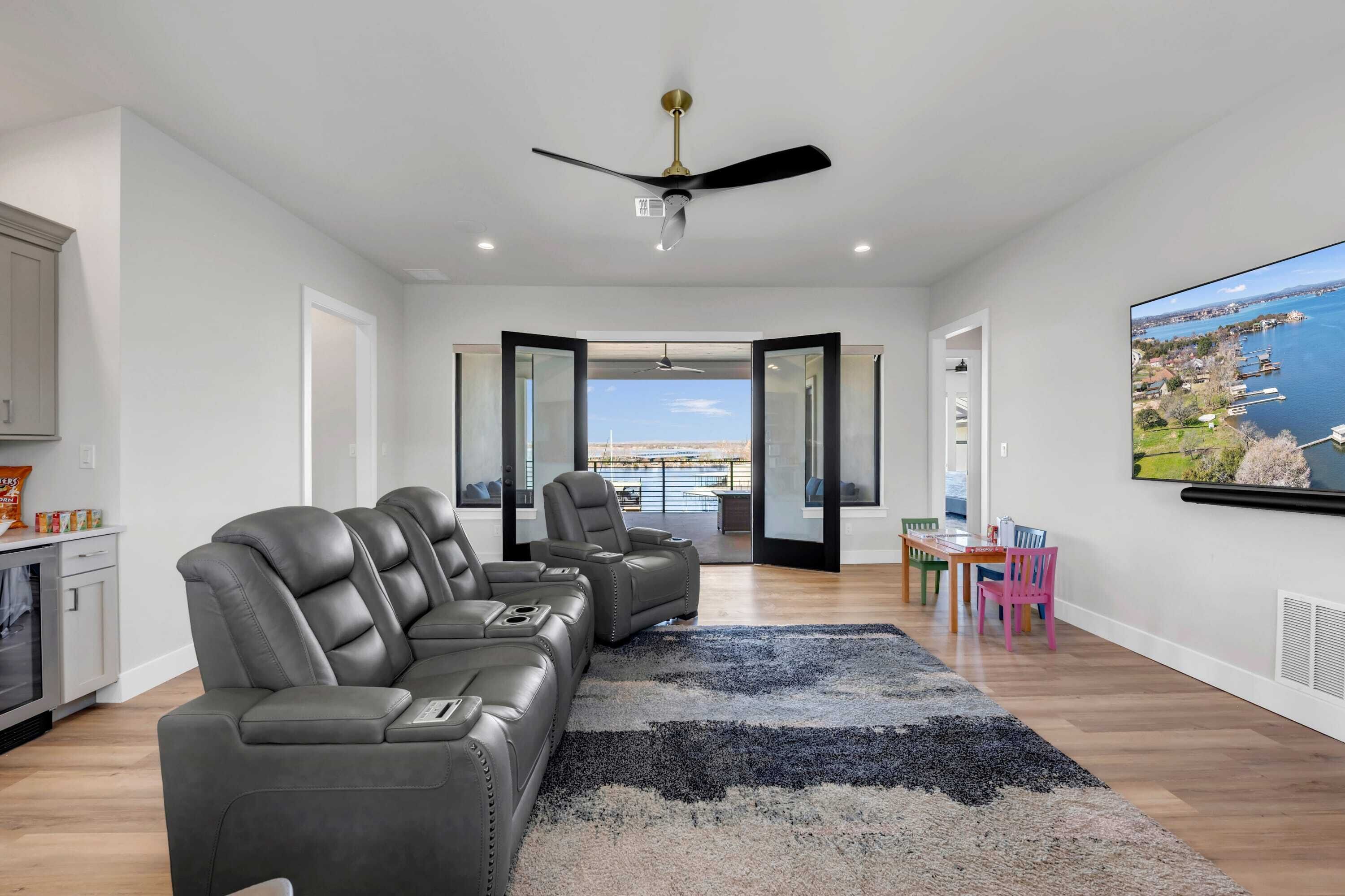 A modern living room with a grey leather sofa, a ceiling fan, a large TV, and a view of a body of water through an open door.