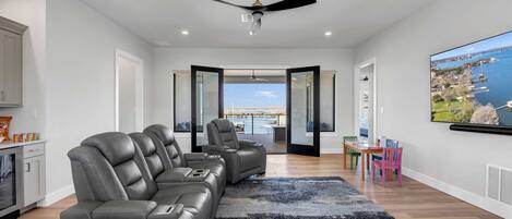 A modern living room with a grey leather sofa, a ceiling fan, a large TV, and a view of a body of water through an open door.