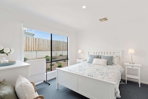 A bedroom with a large window, a bed with a floral bedspread, two bedside tables with lamps, a white dresser, and a chair.