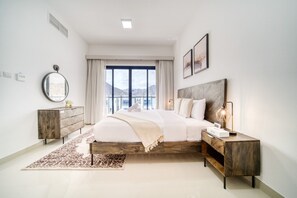 A modern bedroom with a large bed, a wooden nightstand, a dresser, and a view of the outdoors. - Lavish 4BR Villa w/ 2 Asst Rms, Al Dana Island, Fujairah by Deluxe Holiday Homes (Fujairah)