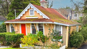 Uma casa charmosa, térrea, com porta vermelha, identificada como “Birdies Cottage”, cercada por vegetação exuberante e um portão branco de piquete.