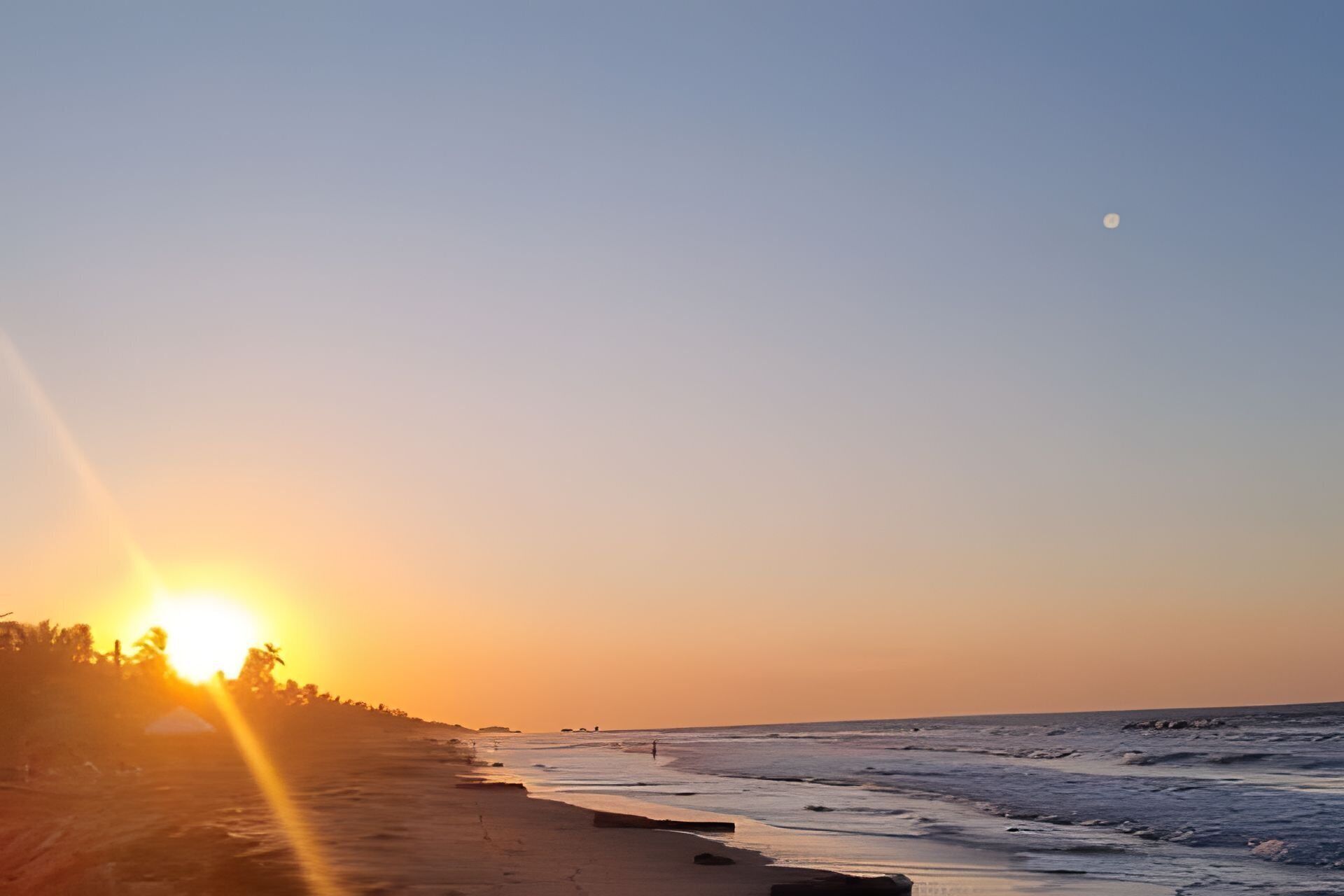 Een strand bij zonsondergang, met de zon laag aan de horizon, golven die zachtjes op het strand komen en een heldere lucht.