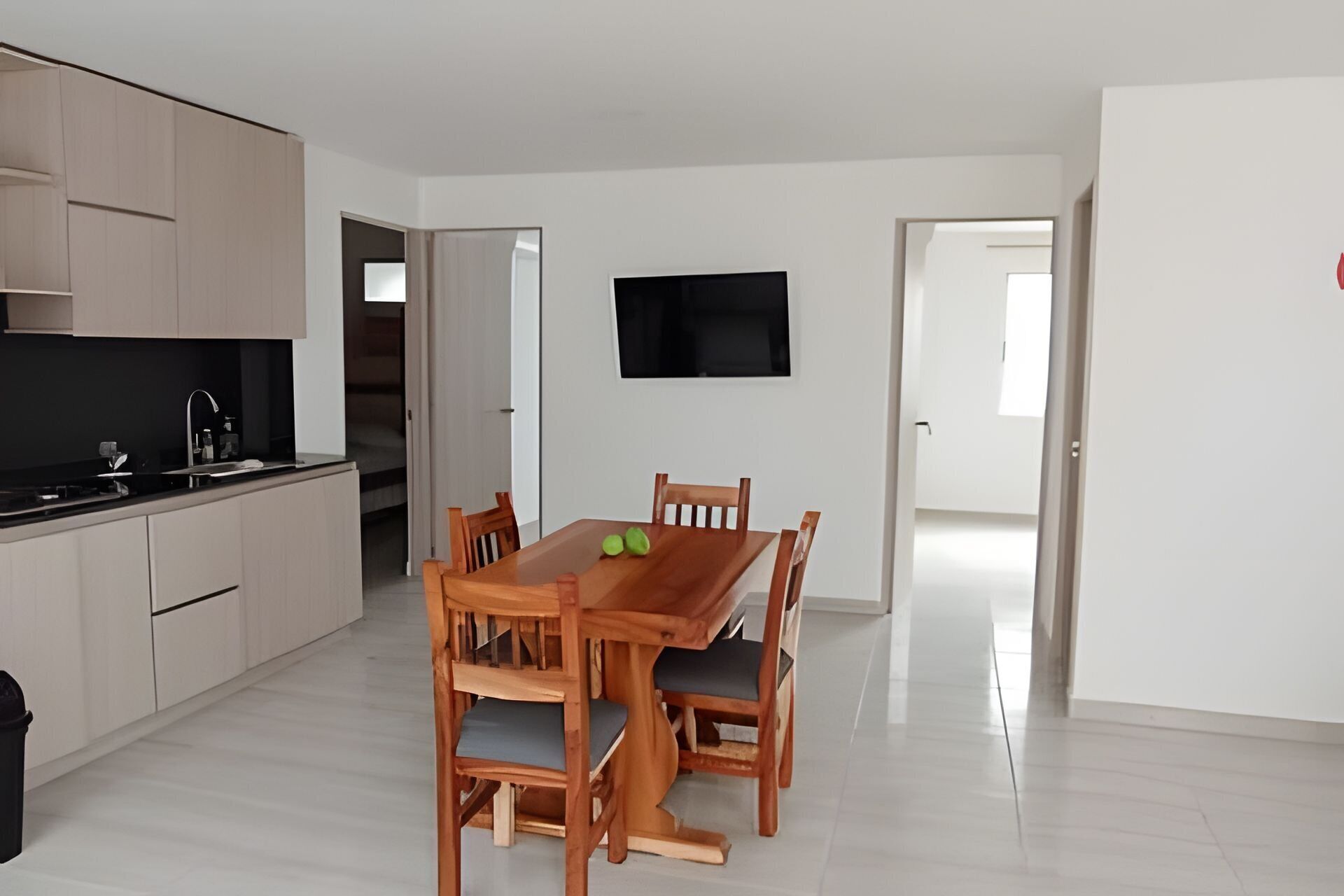 A modern kitchen with light wood cabinets, a black countertop, and a built-in stove. There is a dining area with a wooden table and chairs, and a flat-screen TV mounted on the wall.