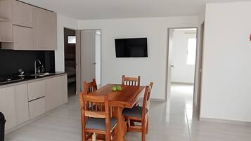 A modern kitchen with light wood cabinets, a black countertop, and a built-in stove. There is a dining area with a wooden table and chairs, and a flat-screen TV mounted on the wall.