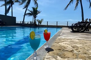 A poolside area with two drinks on a table, palm trees, and a clear blue sky. - APARTAMENTOS EN PALMAR DE LOS VIENTOS  (San Bernardo del Viento)