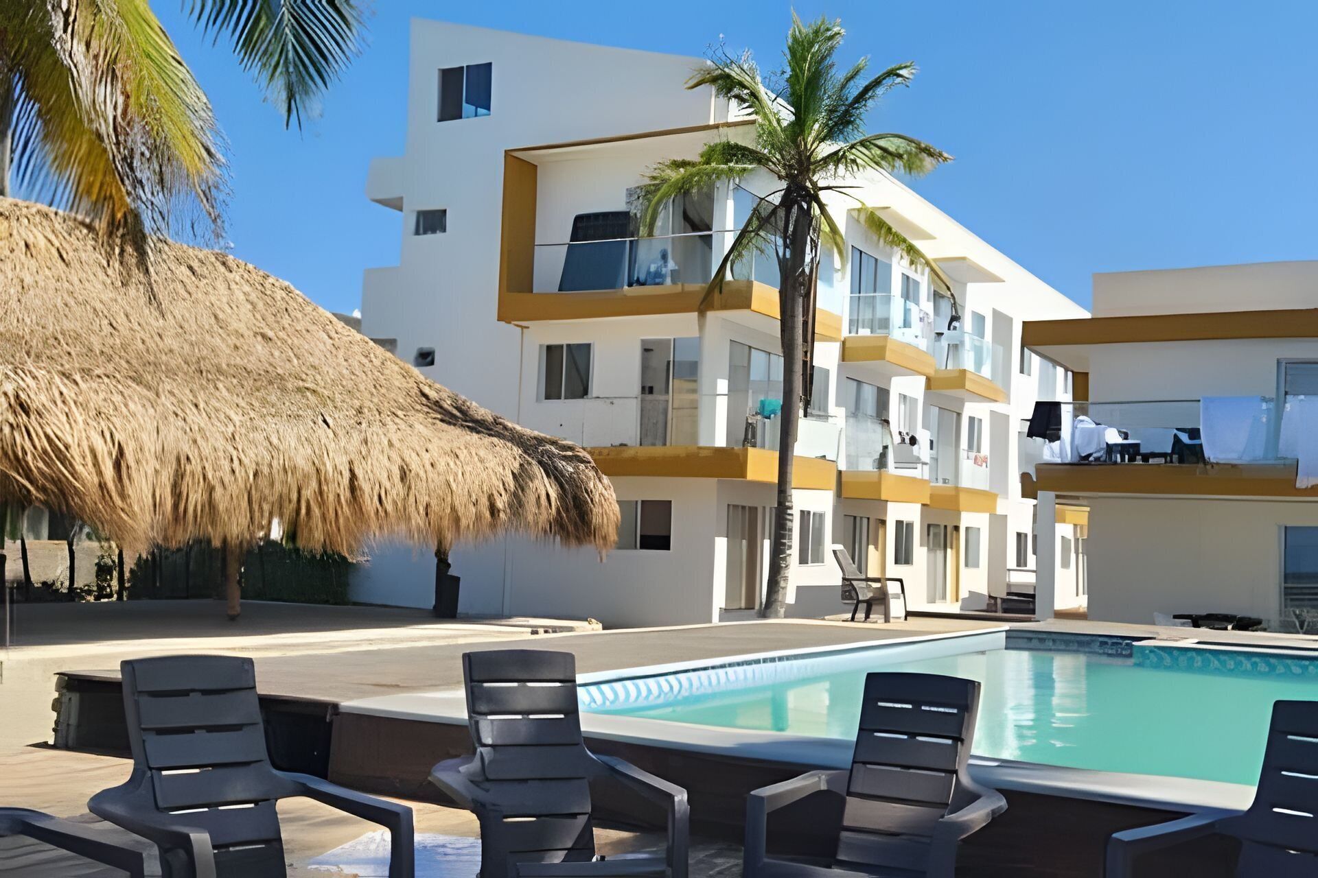 A pool area with lounge chairs, a thatched roof, and a multi-story building with balconies.