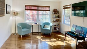 A room with two light blue armchairs, a small table, a floor lamp, a ceiling fan, a bookshelf, and a TV mounted on the wall. - Quiet Mid-Mod Getaway with Hot Tub, Outdoor Seating & Firepit (Asheville)