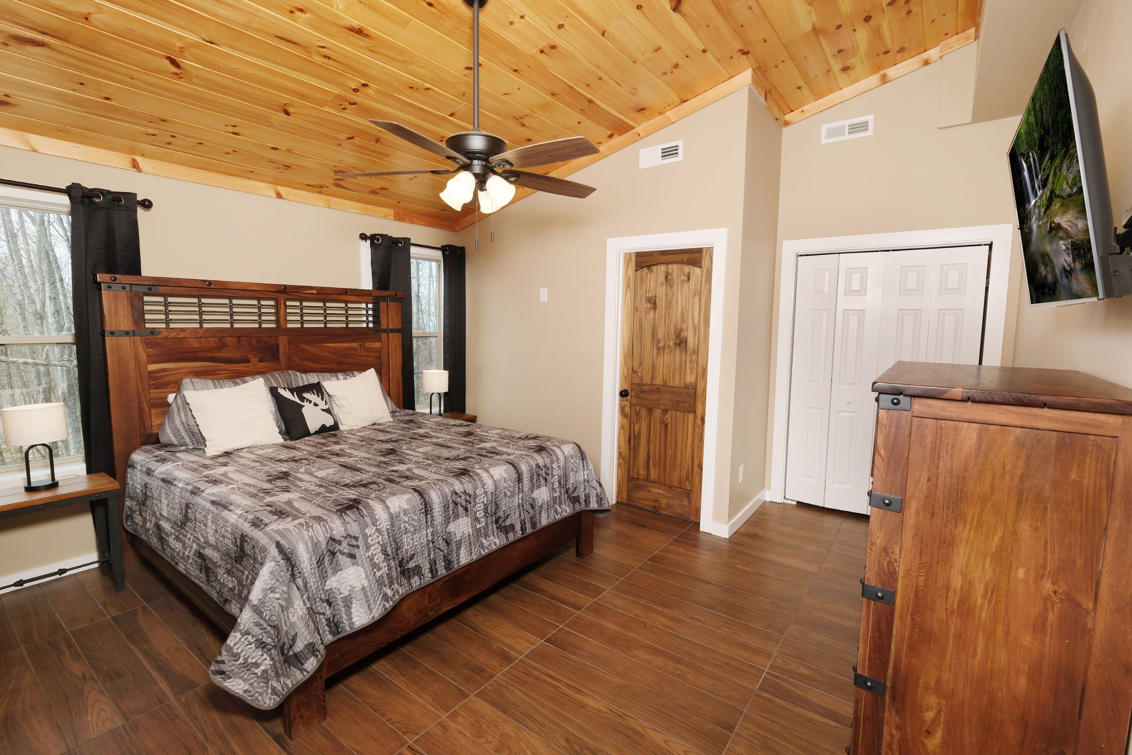 Une chambre avec un plafond en bois, un lit recouvert d’une couvre-lit à motifs, un ventilateur de plafond et un téléviseur fixé au mur.