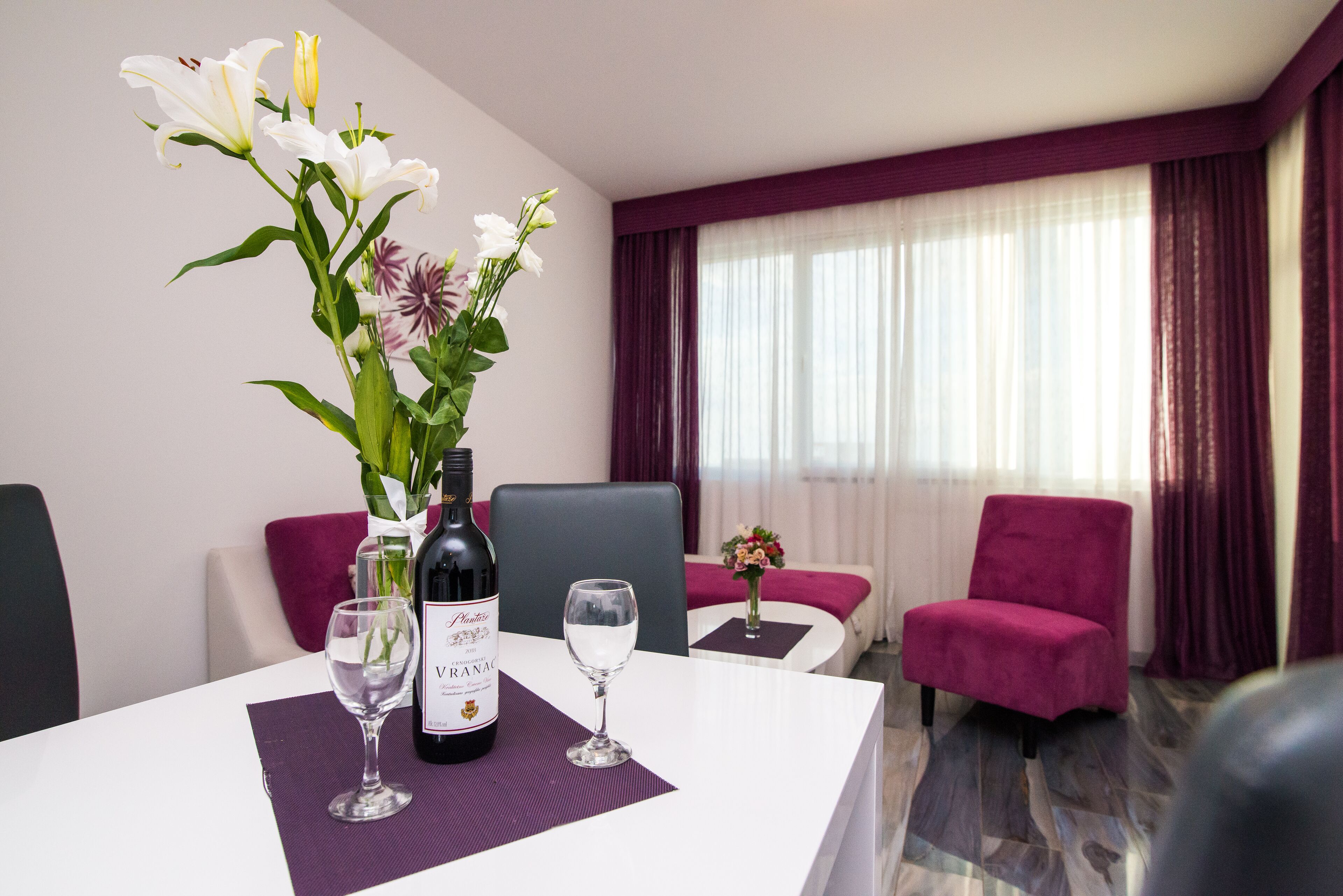 A hotel room with a dining table set with a bottle of wine, two glasses, and a vase of flowers. There is a chair and a small table with a vase of flowers in the corner.