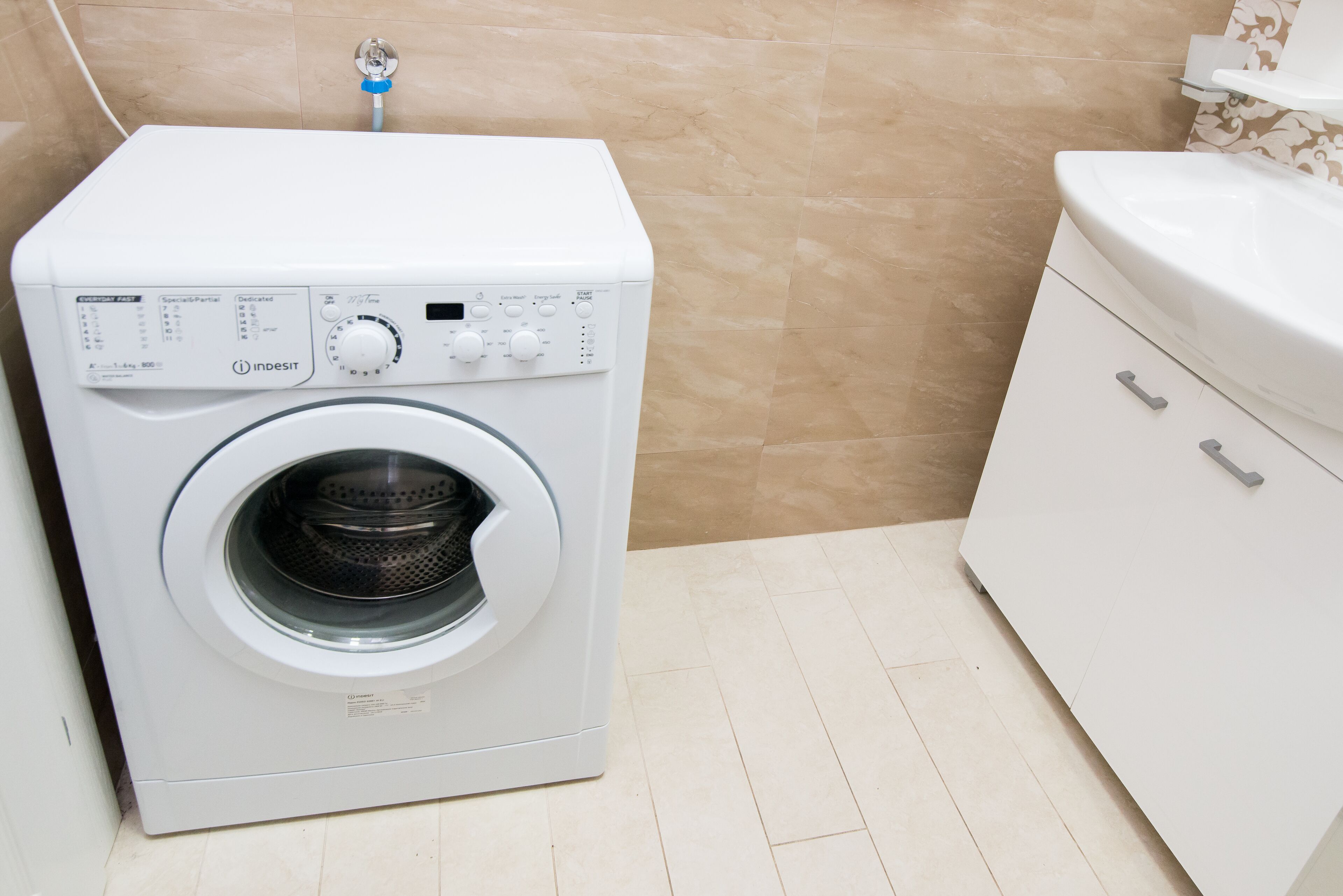 A front-loading washing machine in a bathroom with beige tiles, a white cabinet with drawers, and a white toilet.