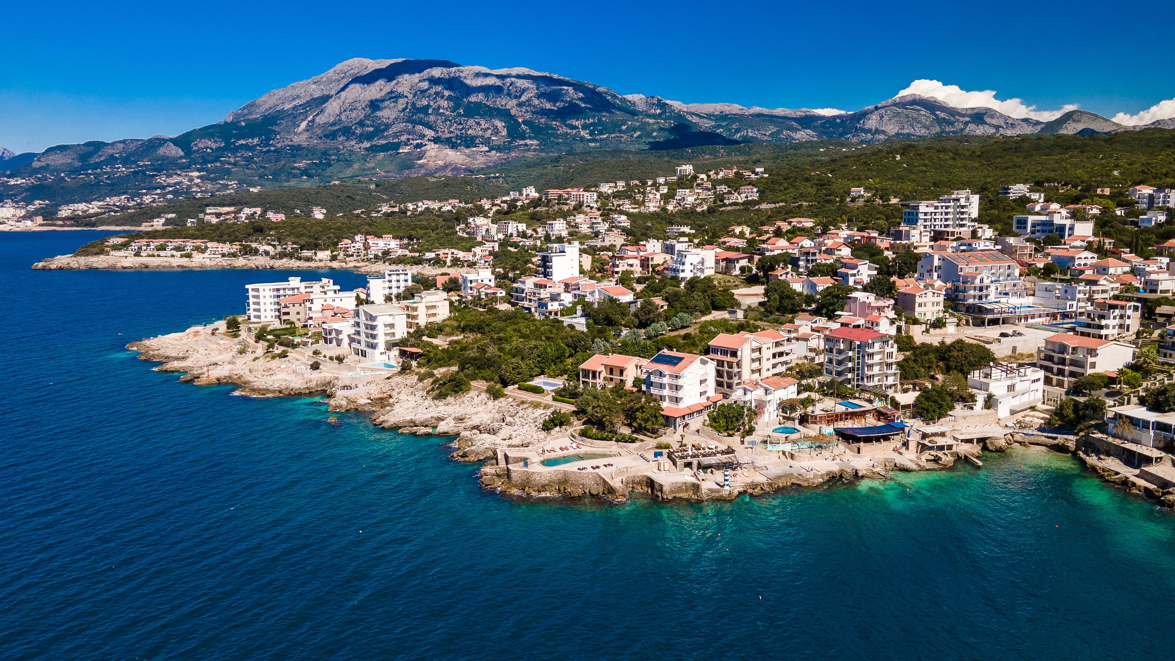 Un pueblo costero con edificios a lo largo de la costa, una montaña en el fondo y aguas azules cristalinas.