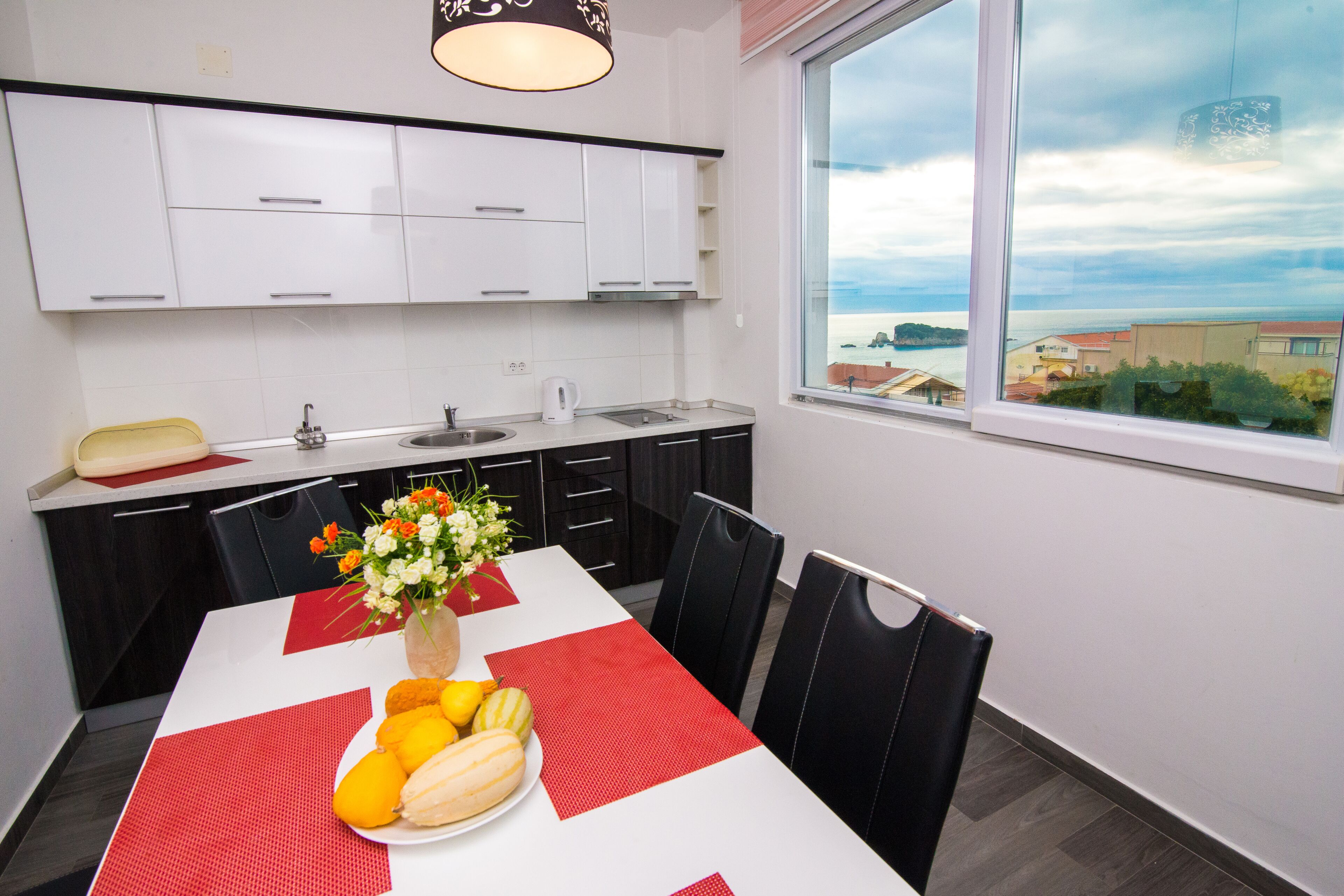 A modern kitchen with white cabinets, a dining table with red placemats, and a view of the sea through a large window.