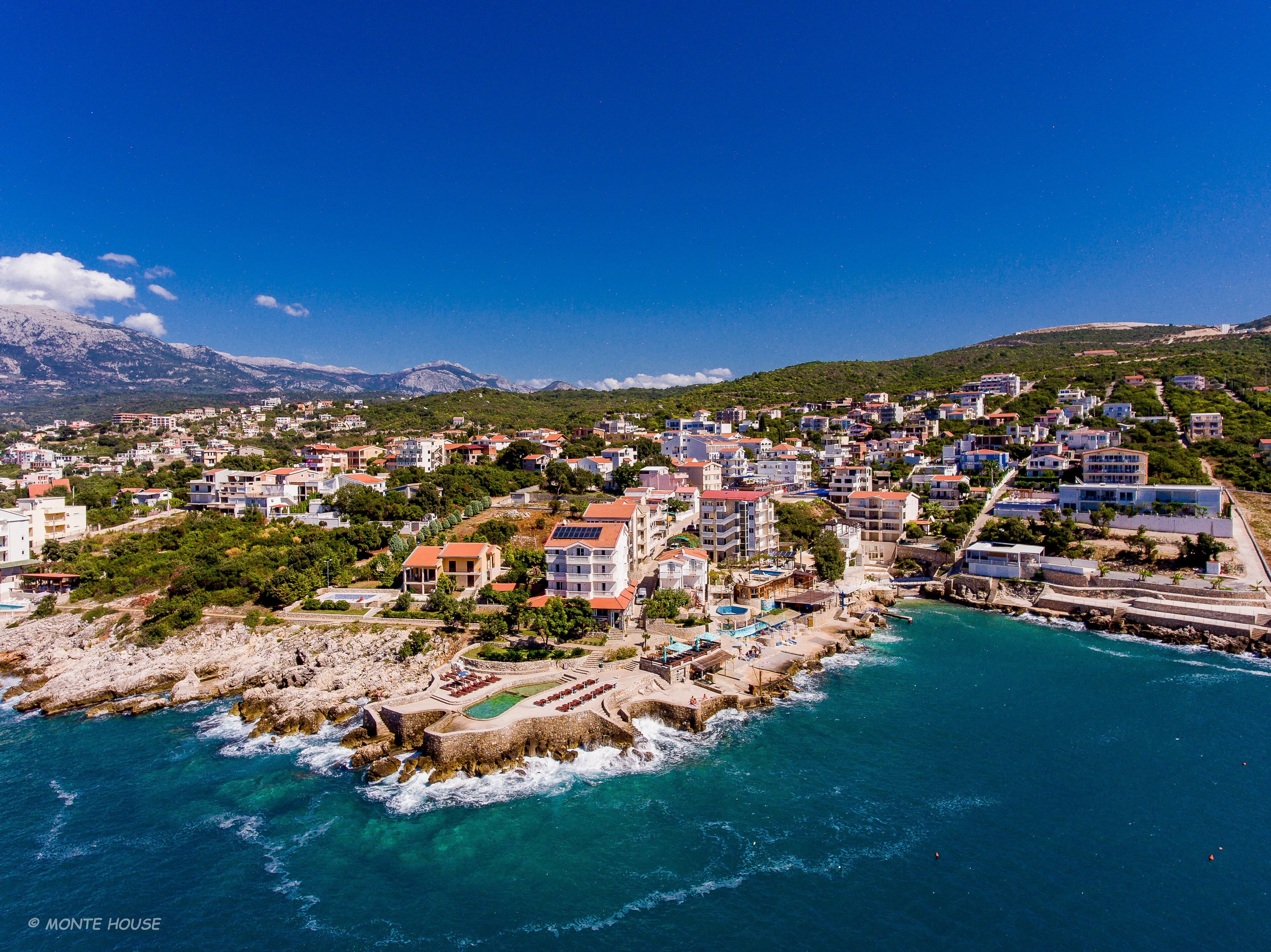 An aerial view of a coastal town with buildings, a rocky shoreline, and a clear blue sky.