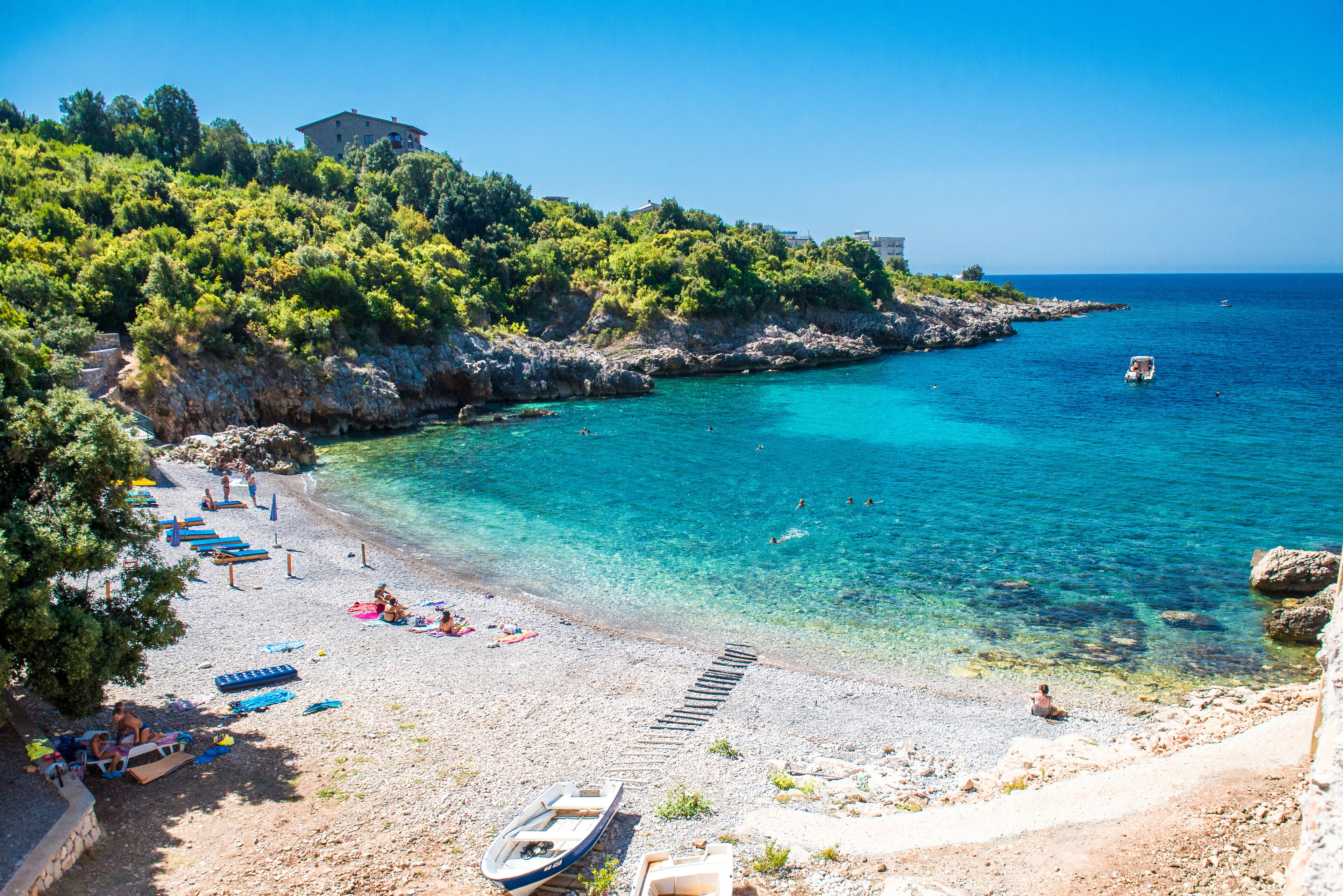 Une plage à l’eau turquoise et limpide, un rivage rocheux et un bâtiment perché sur une colline.