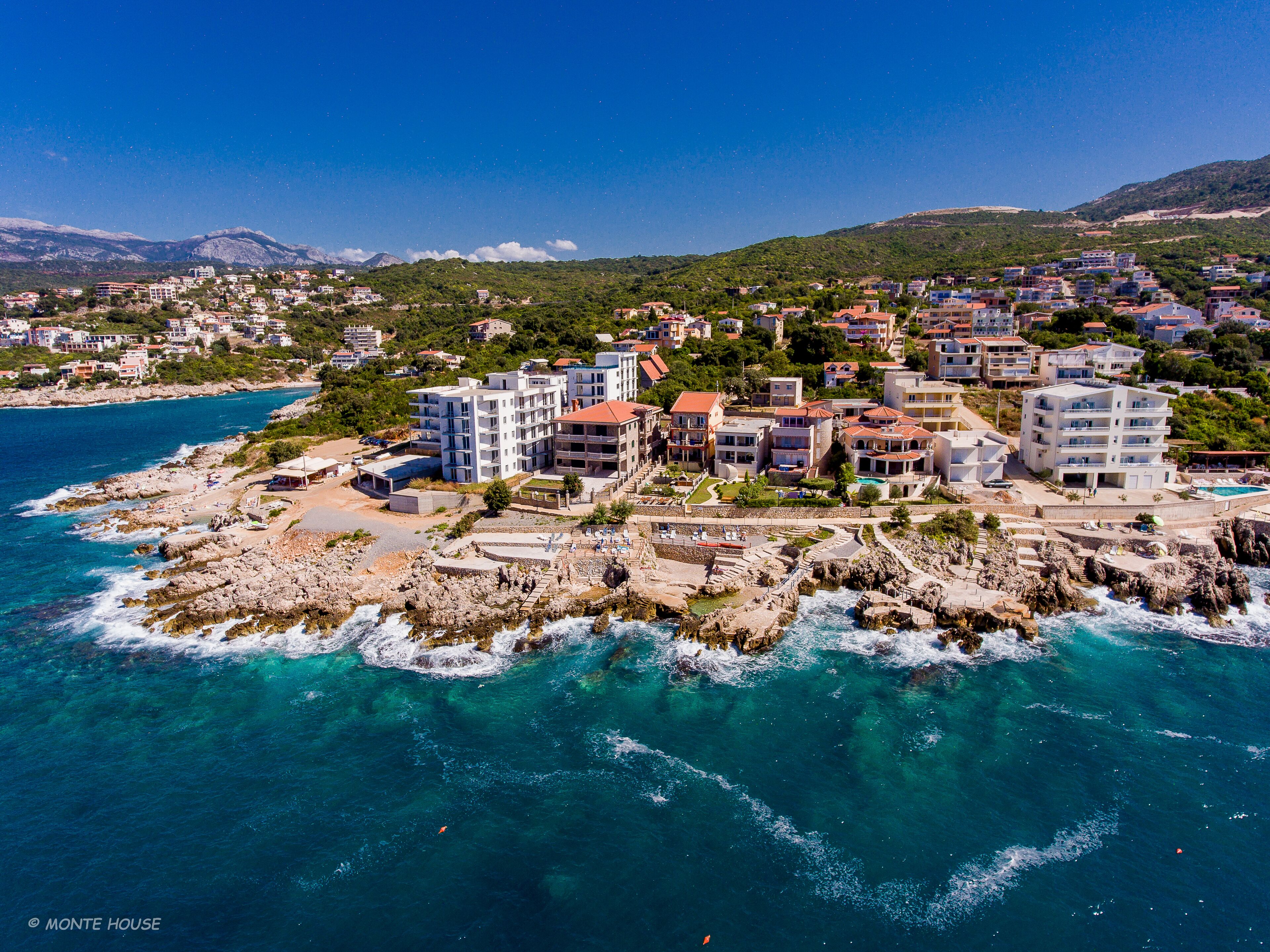 A coastal town with buildings along the shoreline, rocky terrain, and clear blue waters.
