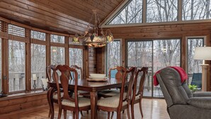 Ein Esszimmer aus Holz mit einem großen Tisch und Stühlen, einer Kristallleuchter und großen Fenstern mit Blick auf Bäume.