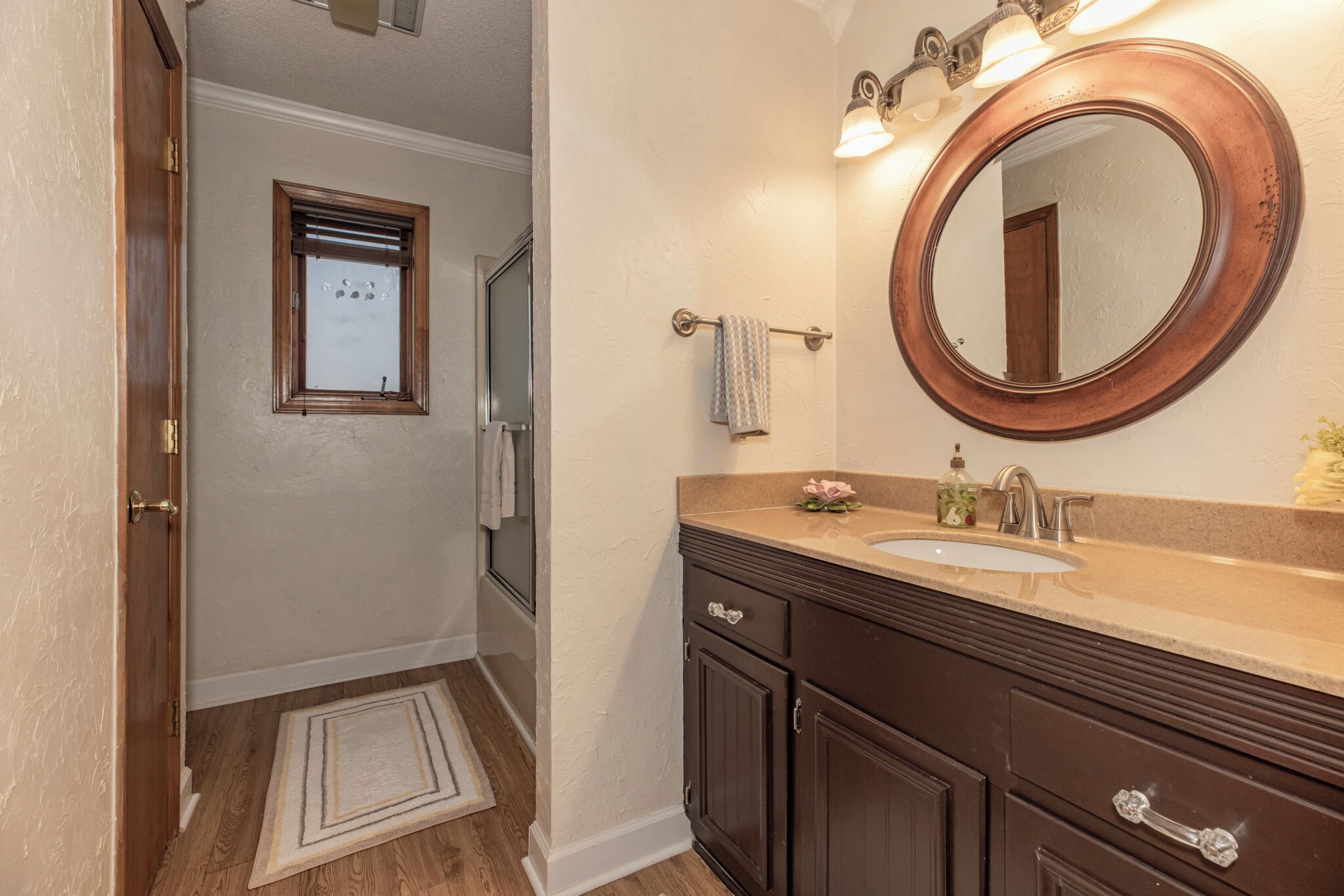A bathroom with a wooden vanity, a round mirror, and a window with blinds.