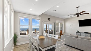 A spacious living area with a dining table, white chairs, a gray sofa, and a ceiling fan.