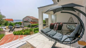 A balcony with a hanging chair and a table, overlooking a street and a body of water.