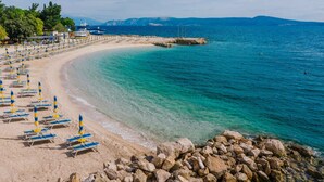 A beach with rows of sun loungers and umbrellas, a rocky shoreline, and a clear view of the sea and distant mountains.