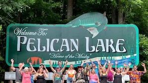 Un grupo de personas frente a un cartel de bienvenida a Lago Pelican, con un gráfico de un pez grande y árboles de fondo.