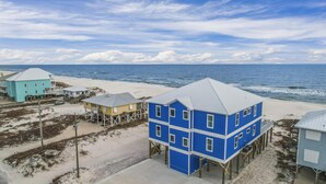 Ett kustnära område med flera hus som ligger direkt vid stranden, inklusive ett framträdande blått hus med ett sadeltak, som är byggt på pålar över sanden.