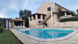 Una casa de dos pisos con piscina, mobiliario exterior y un patio cubierto.
