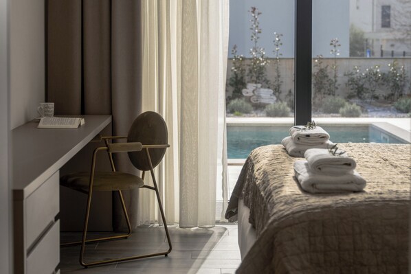 A bedroom with a bed, a desk, and a chair. There is a window with a view of a pool and greenery.