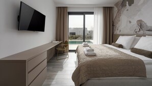 Ett modernt hotellrum med en platt-TV, ett skrivbord, en stol, en säng med ett beige täcke och utsikt över en swimmingpool och byggnader.