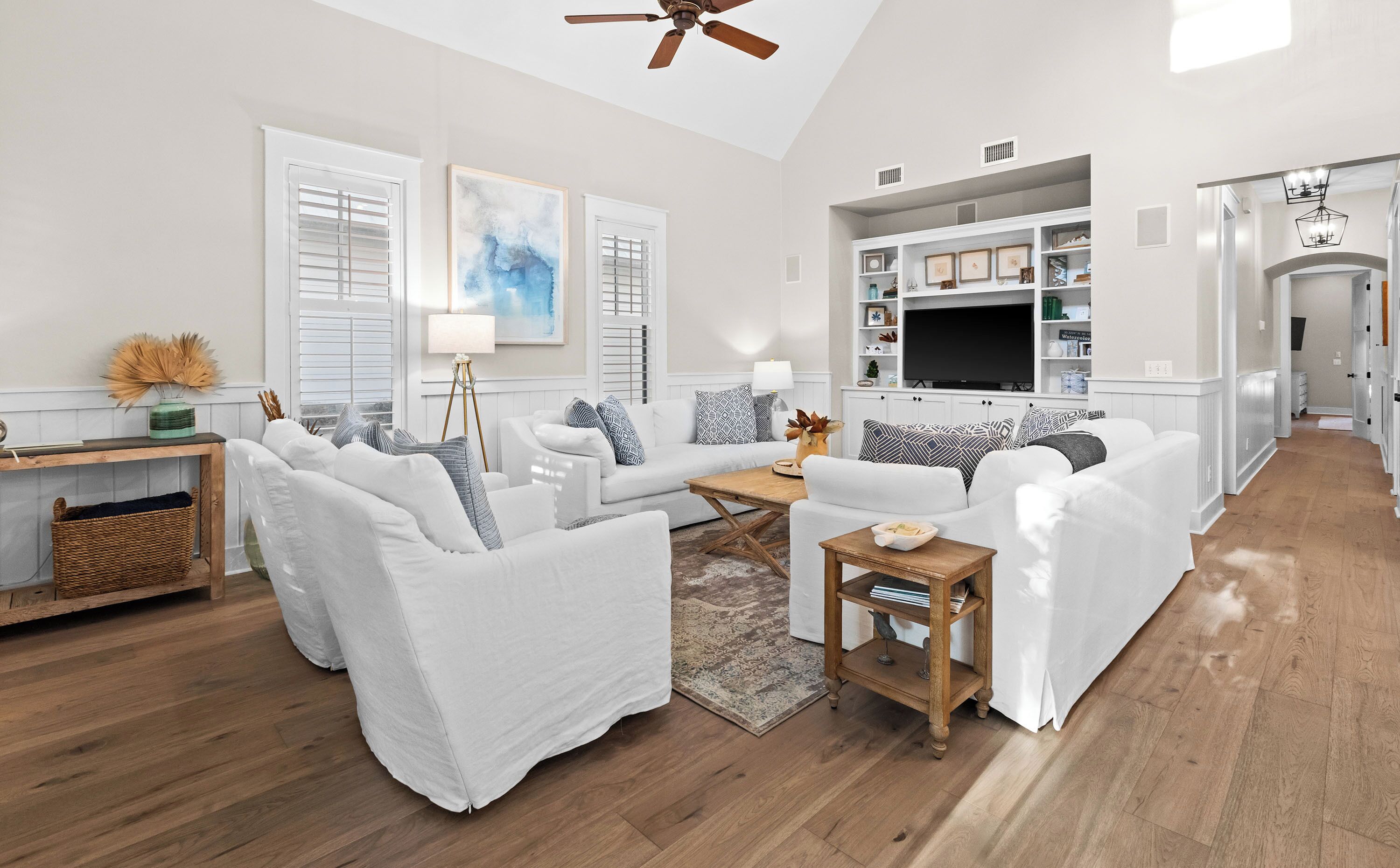 A spacious living room with a large sofa set, a coffee table, and a TV. There is a dining area with a wooden table and chairs. The room has white walls and wooden flooring.