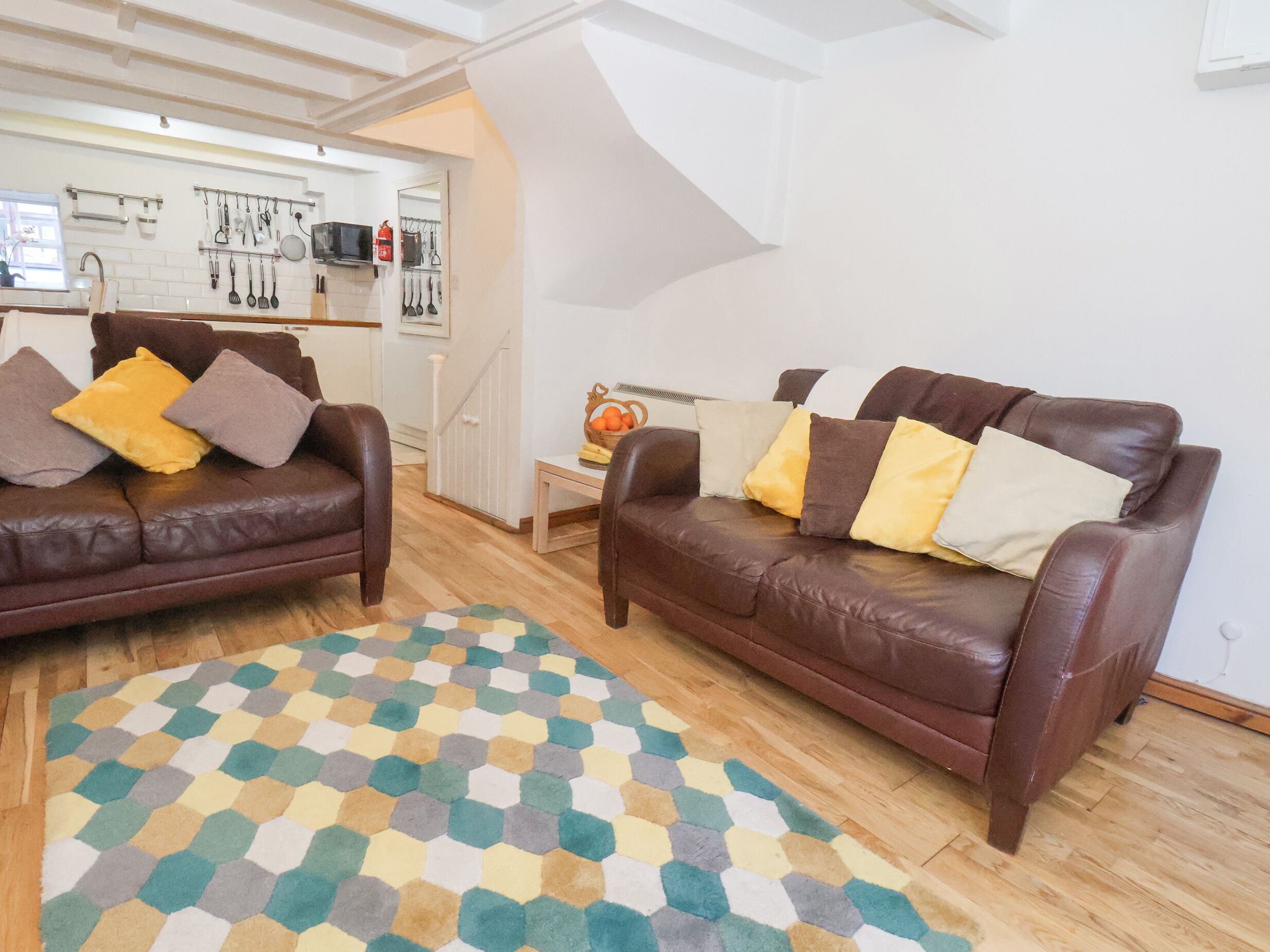 Una sala de estar con dos sofás de cuero marrón, un tapete hexagonal de colores y una zona de cocina al fondo.
