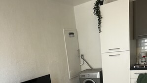 Kompakti oleskelutila, jossa on valkoinen, litteänäyttöinen televisio, pyykinpesukone, pieni keittokomero, jossa on kahvinkeitin, sekä pieni pöytä ja kulho.