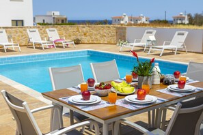 Un espace repas aménagé au bord de la piscine, avec une table pour deux, entouré de chaises longues, d’un mur en pierre et offrant une vue sur la mer.