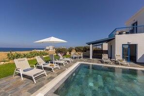 Un espace piscine avec des chaises longues et un parasol blanc, donnant sur une plage.