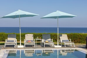 Un espace aménagé au bord de la piscine, avec des chaises longues et des parasols, une petite table avec une boisson et des fruits, et une vue sur la mer.