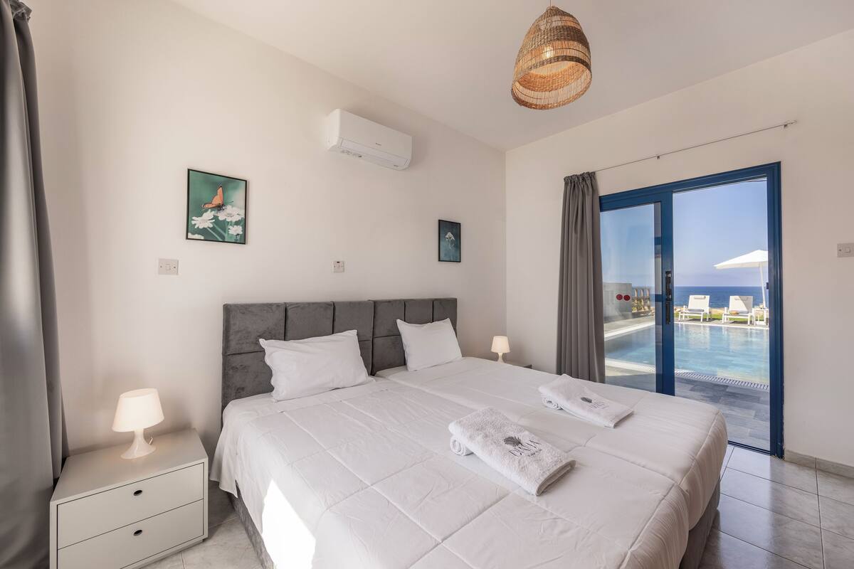 Quarto de hotel com cama, mesa de cabeceira, luminária, ar condicionado, quadro na parede e vista para uma piscina através de uma porta de vidro.