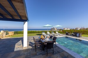 A poolside area with a table and chairs, umbrellas, and a view of the sea. - Diana Seaside Villas (Pomos)