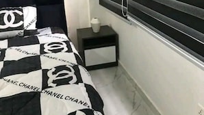 Ett sovrum med en säng som har ett svart- och vitrutigt överkast med Chanels logotyp, ett sängbord med en lampa och ett fönster med persienner.