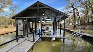 A covered dock with a boat moored at the end, surrounded by water and trees. - Richland Chambers Lakefront 2-story home with boat dock and pavilions. (Streetman)