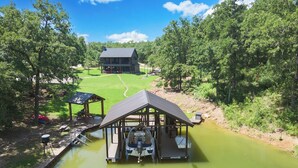 A riverside cabin with a dock, surrounded by lush greenery and a large house in the background. - Richland Chambers Lakefront 2-story home with boat dock and pavilions. (Streetman)