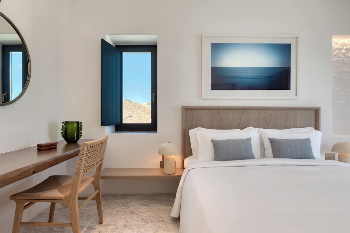 A modern bedroom with a bed, a desk, a chair, a window with a view, a framed picture on the wall, and a bedside table with a lamp.