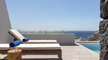 A sunlit outdoor lounge area with two lounge chairs, a wooden stool, and a glass of water.