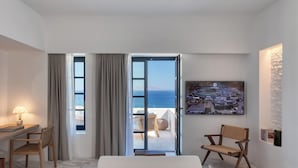 A hotel room with a bed, a desk, a chair, a view of the sea, and a painting of the hotel.