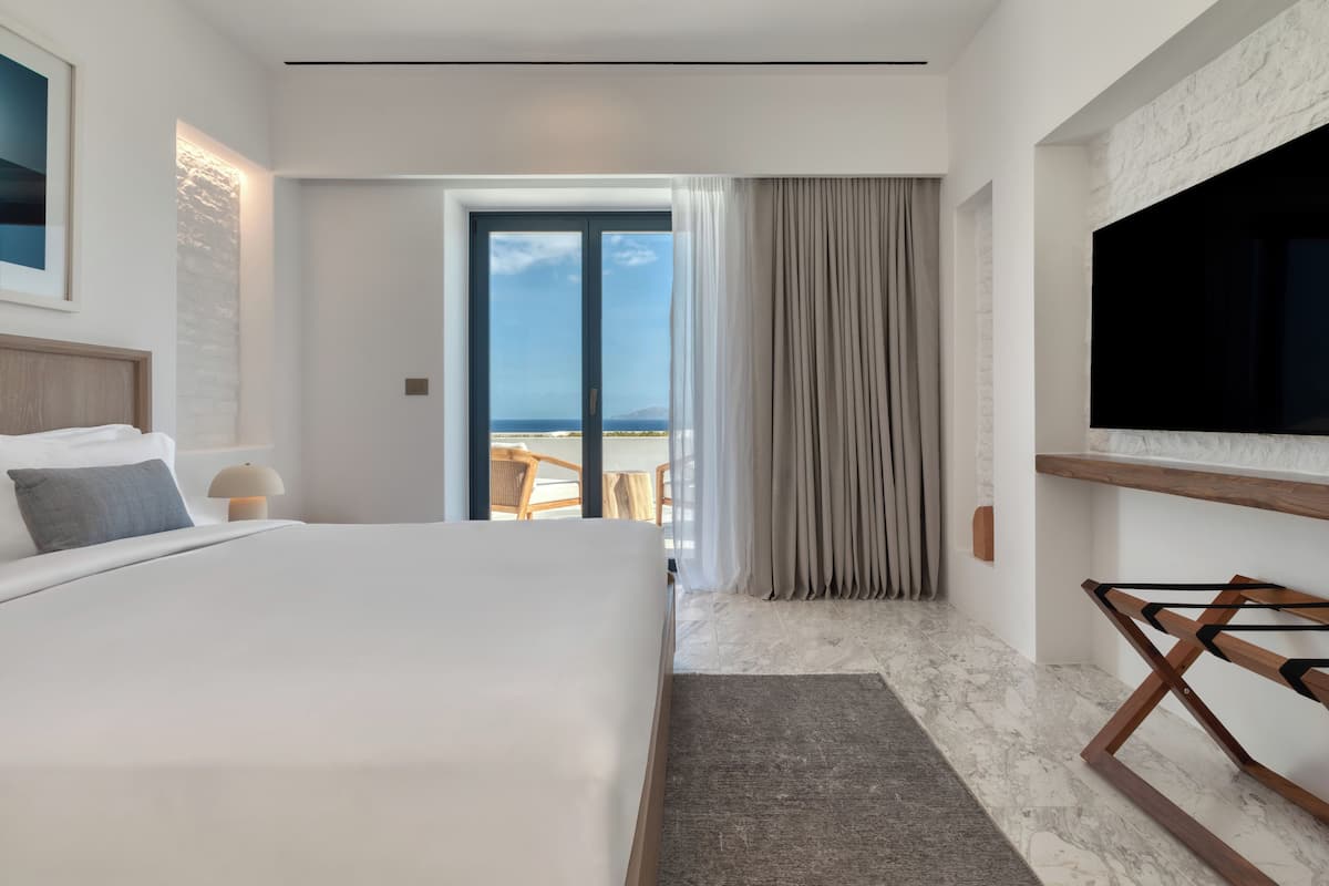 A modern hotel room with a large bed, a TV mounted on the wall, and a view of the ocean through a sliding door.